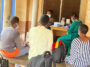 Trois personnes sont assises sur un banc et nous tournent le dos. En arrière-plan, un professionnel de la santé portant une blouse verte, un masque facial et des clous de girofle violets semble préparer un échantillon. L'équipement est posé sur la table où elle est assise et on peut voir une femme assise sur une chaise à côté de la table.