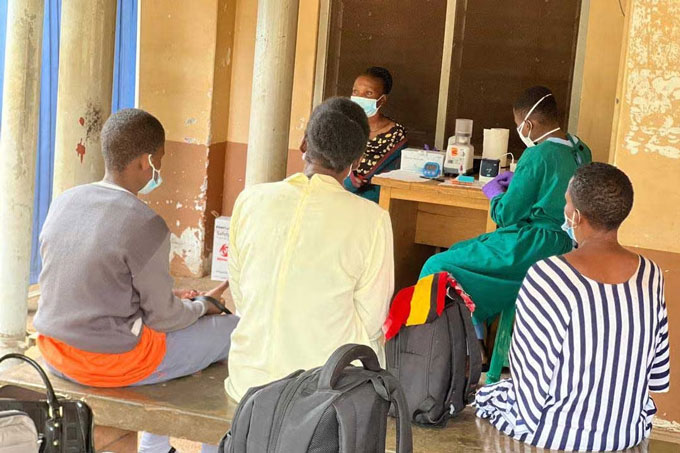 Trois personnes sont assises sur un banc et nous tournent le dos. En arrière-plan, un professionnel de la santé portant une blouse verte, un masque facial et des clous de girofle violets semble préparer un échantillon. L'équipement est posé sur la table où elle est assise et on peut voir une femme assise sur une chaise à côté de la table.
