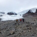 Trois personnes en tenue de temps froid traversent une pente parsemée de rochers, avec des glaciers enneigés visibles derrière eux.