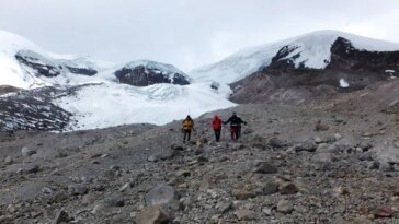 Trois personnes en tenue de temps froid traversent une pente parsemée de rochers, avec des glaciers enneigés visibles derrière eux.