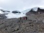 Trois personnes en tenue de temps froid traversent une pente parsemée de rochers, avec des glaciers enneigés visibles derrière eux.
