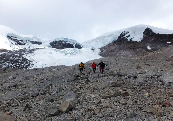 Trois personnes en tenue de temps froid traversent une pente parsemée de rochers, avec des glaciers enneigés visibles derrière eux.