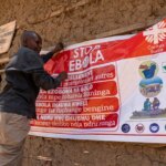 Une personne tend une grande affiche qui dit "ARRÊTEZ EBOLA" sur le côté d'un bâtiment construit en terre crue