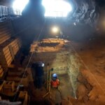 Un chercheur casqué se tient dans une immense fosse creusée dans le sol d'une grotte, entouré d'équipements archéologiques.