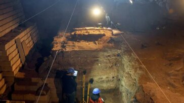 Un chercheur casqué se tient dans une immense fosse creusée dans le sol d'une grotte, entouré d'équipements archéologiques.
