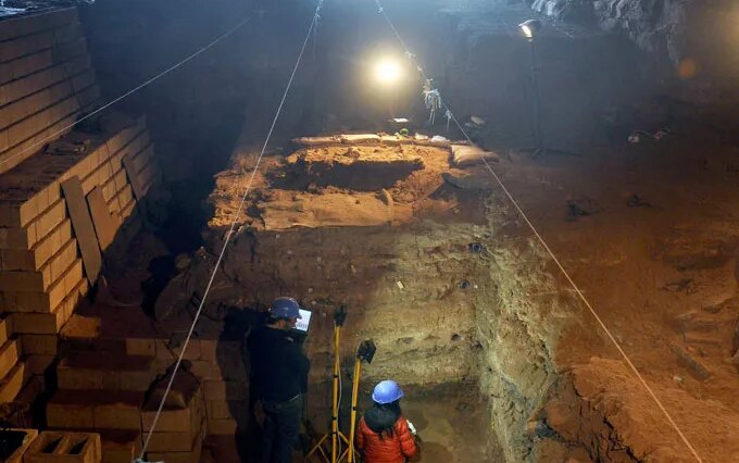 Un chercheur casqué se tient dans une immense fosse creusée dans le sol d'une grotte, entouré d'équipements archéologiques.