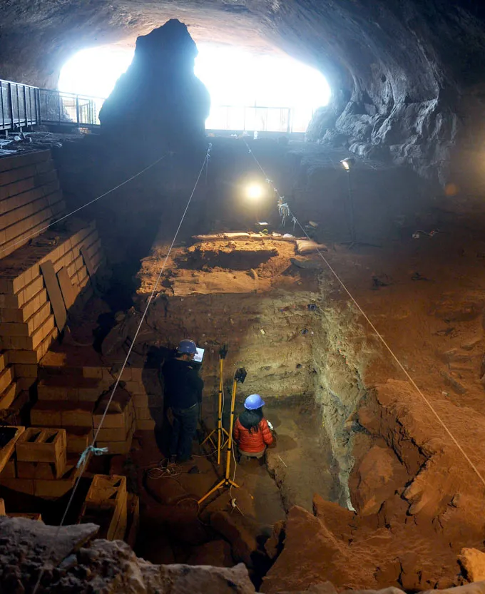Un chercheur casqué se tient dans une immense fosse creusée dans le sol d'une grotte, entouré d'équipements archéologiques.