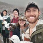 Quatre personnes sourient sur une plage rocheuse, brumeuse et froide. Ils tiennent des mouettes en plastique.