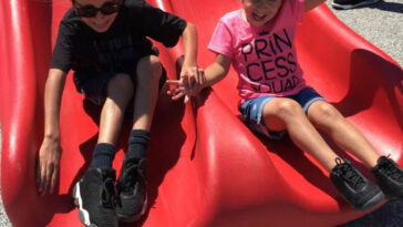 Deux enfants descendent un toboggan rouge en se tenant la main.