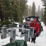 Les travaux d'hiver montrent qu'il est vraiment payant d'être une mésange des montagnes intelligente Dans une forêt enneigée, une personne vêtue d'un parka rouge examine un ensemble de huit mangeoires à oiseaux attachées à un système de poulies pour les hisser hors de portée des ours.