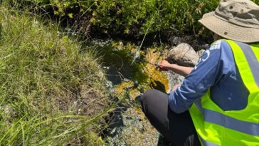 Une personne portant un gilet jaune vif s’accroupit près d’une source chaude pour prélever un échantillon d’eau.