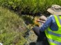 Une personne portant un gilet jaune vif s’accroupit près d’une source chaude pour prélever un échantillon d’eau.